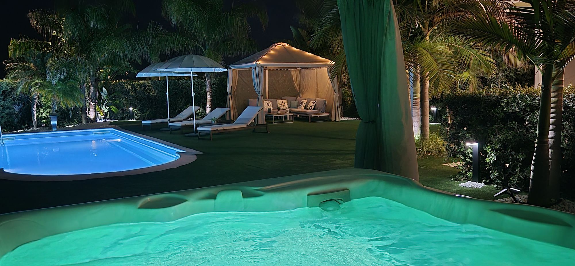 Jacuzzi illuminata di turchese di sera, con piscina e gazebo sullo sfondo